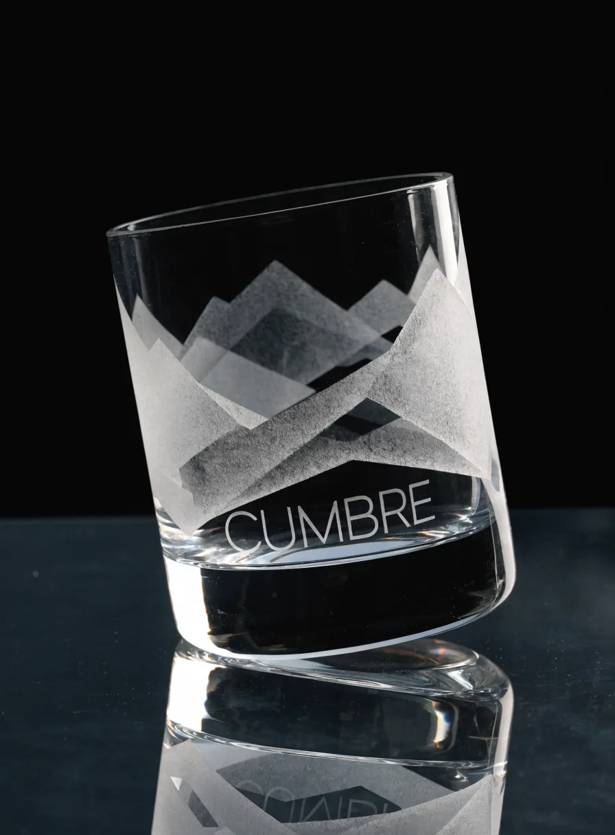 vaso bajo de cristal grueso sobre una superficie negra reflejante, con una sierra de montañas arenada en planos de distinta densidad y el nombre CUMBRE en letras claras debajo del dibujo