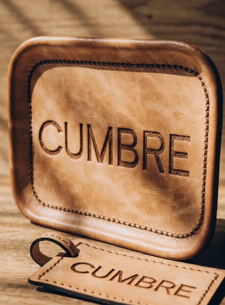 charola y llavero de piel curtida grabados a láser con el nombre CUMBRE hundido en el cuero, sobre una mesa de madera