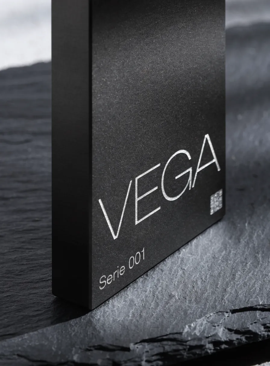 placa cuadrada de aluminio anodizado negro de pie sobre una laja de pizarra oscura, marcada a láser en plata brillante con el nombre VEGA, la leyenda Serie 001 y un código cuadrado en la esquina