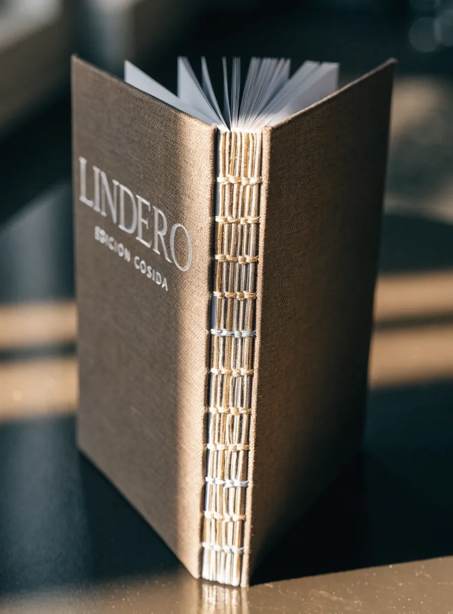 libro entelado en color arena de la marca ficticia LINDERO abierto en ángulo para mostrar el lomo cosido a mano con hilo a la vista, con el título en la portada