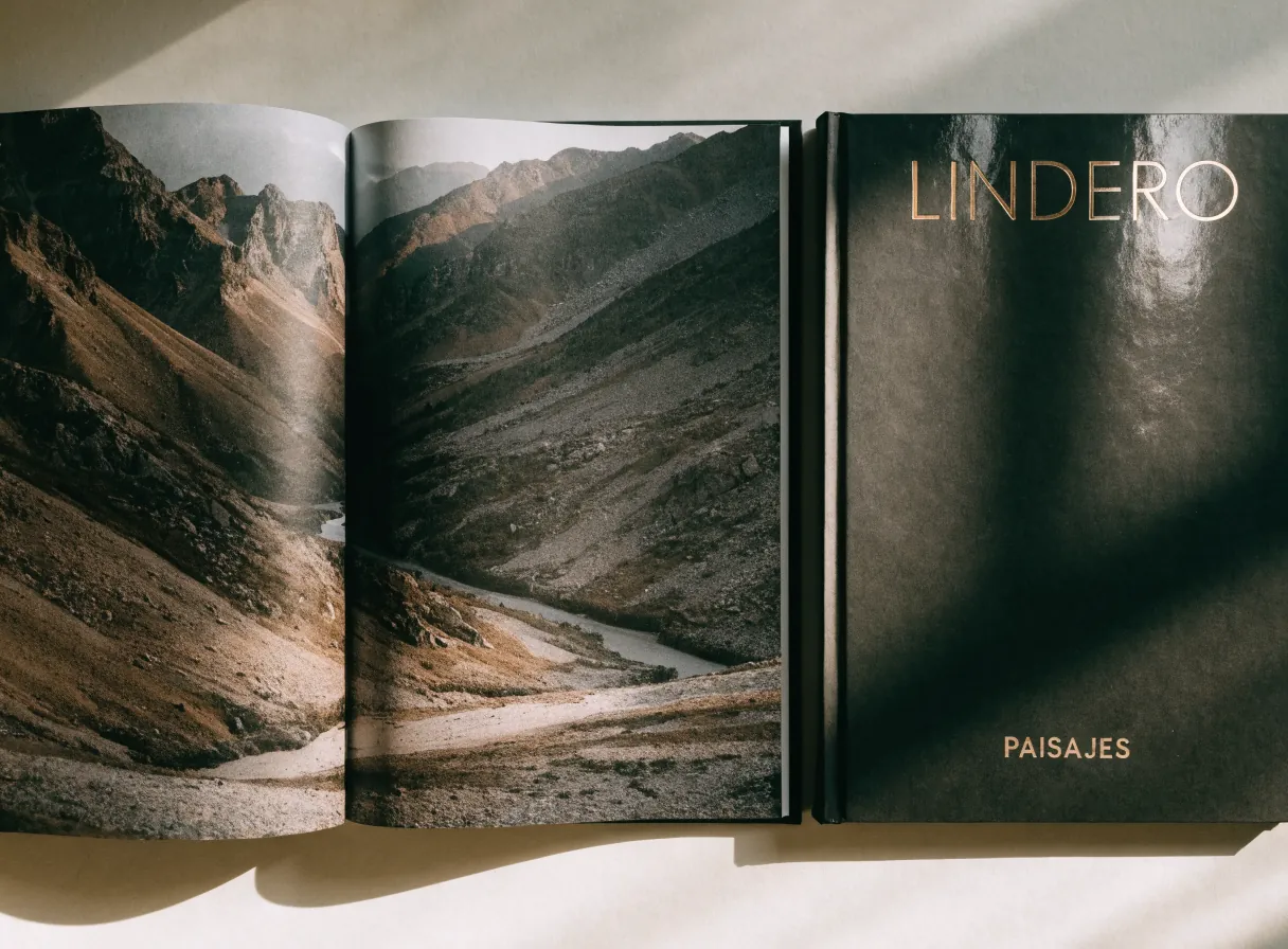 fotolibro de tapa dura de la marca ficticia LINDERO abierto en un paisaje de montaña a doble página, junto al libro cerrado con LINDERO en foil dorado y la palabra PAISAJES