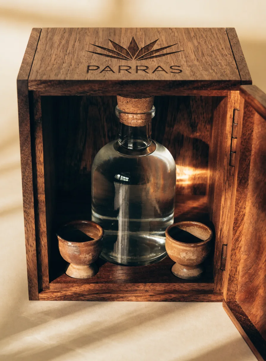 estuche de nogal abierto con una botella de mezcal y dos copitas de barro adentro, la tapa grabada con un agave y la marca PARRAS