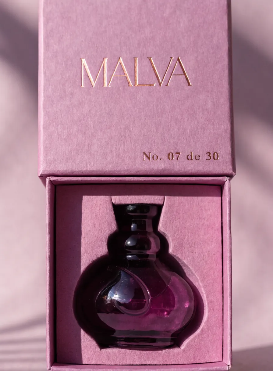 caja de presentación color malva abierta con la marca MALVA en foil oro y No. 07 de 30, un frasco de vidrio morado descansando en el troquel a la medida del interior