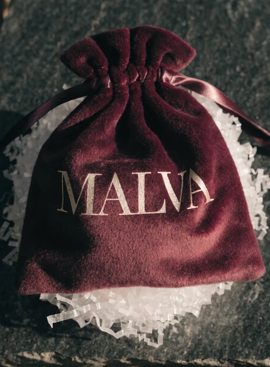 bolsa de terciopelo vino con cordón de satín y la marca MALVA en foil oro, sobre relleno de papel blanco picado