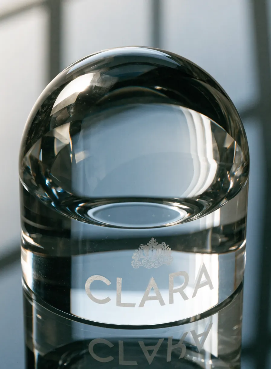 pisapapeles de cristal óptico macizo con la marca CLARA y un escudo grabados al ácido en la base, con luz de ventana entrando por el vidrio