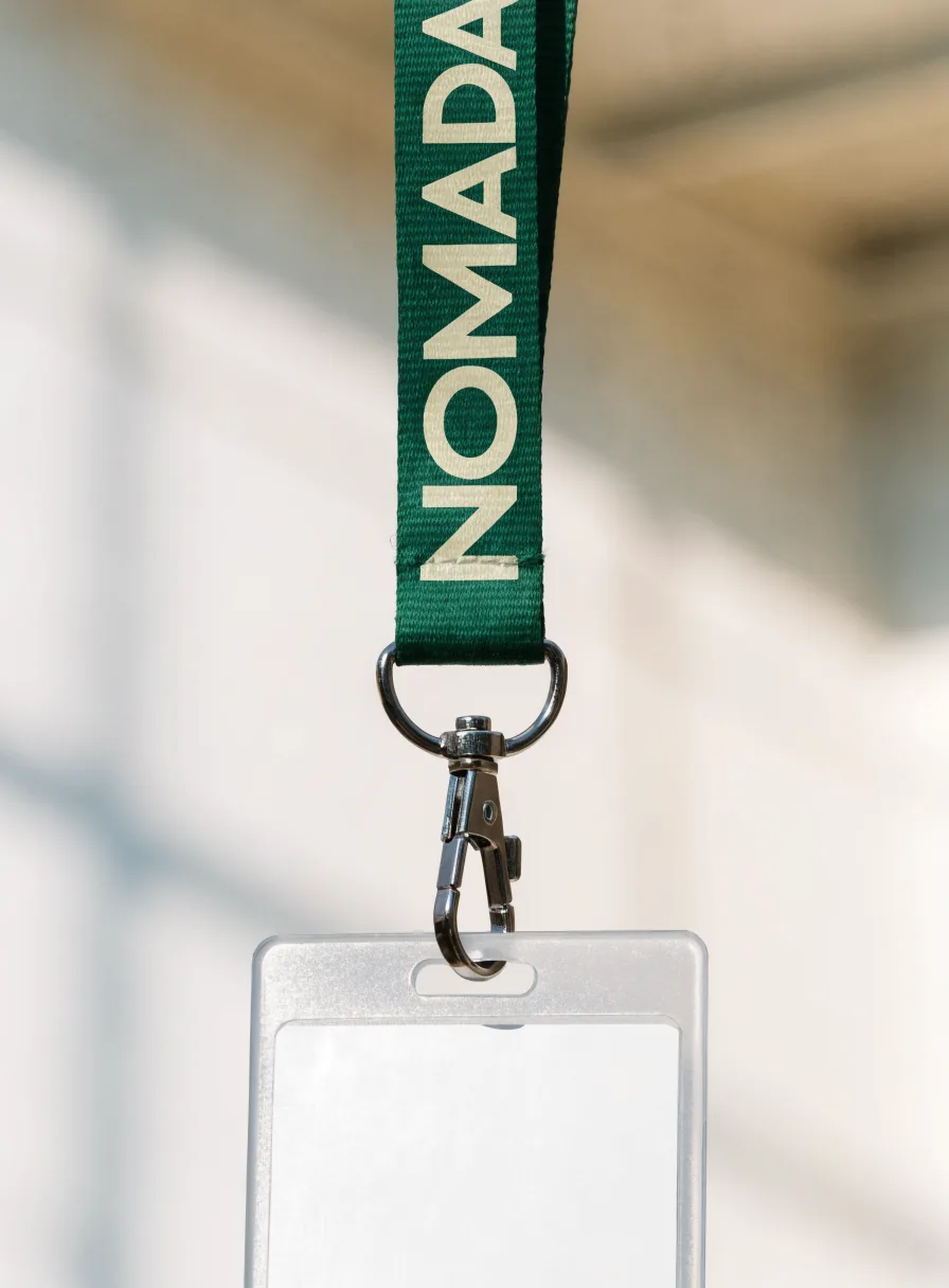 lanyard de cinta verde con la marca Nómada impresa en crema, con broche giratorio de metal y un porta-gafete transparente colgando