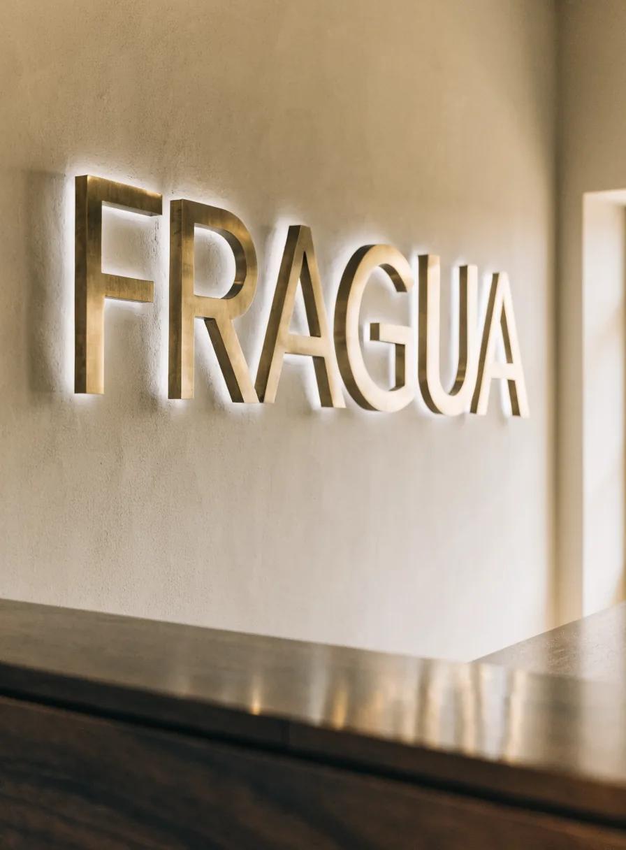 letras corpóreas de latón con la marca ficticia FRAGUA instaladas y retroiluminadas sobre un muro de yeso, encima de un mostrador de recepción