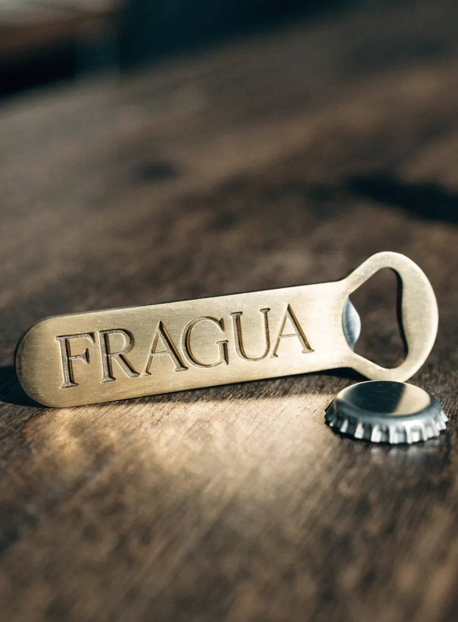 destapador de latón cepillado con la marca ficticia FRAGUA grabada en relieve, junto a una corcholata, sobre una mesa de madera