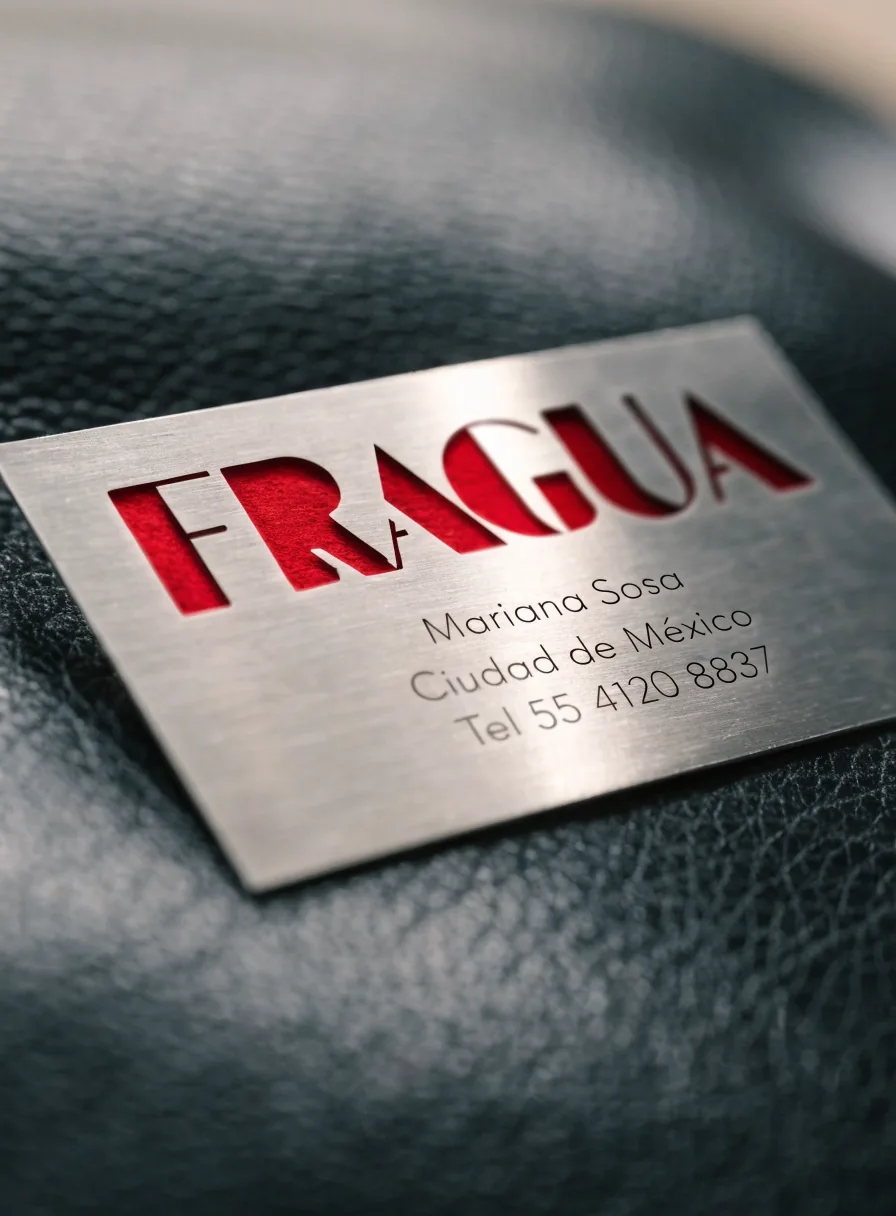 tarjeta de presentación de acero inoxidable con la marca ficticia FRAGUA calada dejando ver un fondo rojo, y el nombre, la ciudad y el teléfono de una persona ficticia grabados, sobre piel negra