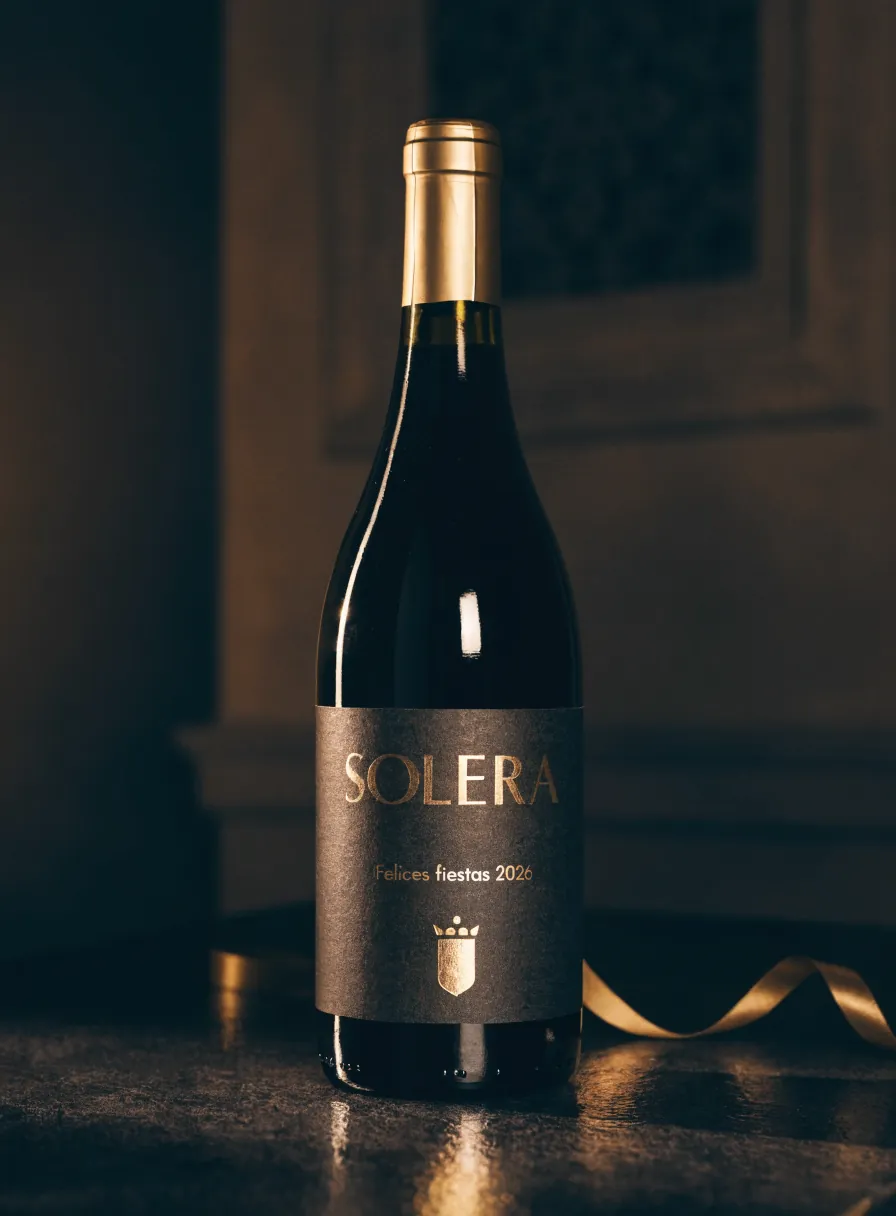 botella de vino tinto con etiqueta de temporada de la marca ficticia Solera en foil dorado, Felices fiestas 2026, con capuchón dorado sobre una mesa oscura con listón dorado
