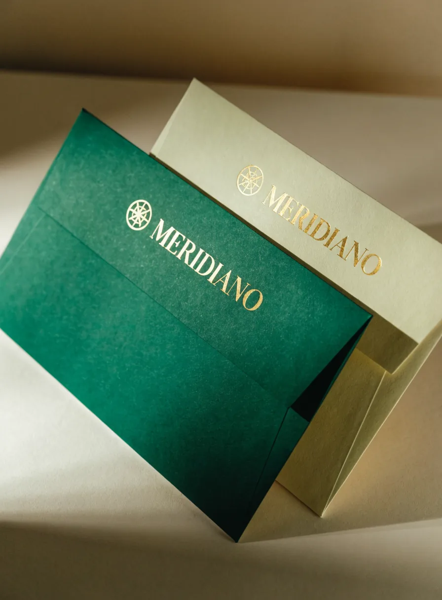 dos sobres de la marca Meridiano, uno verde y uno crema, con el mismo emblema en foil dorado, apoyados uno sobre otro