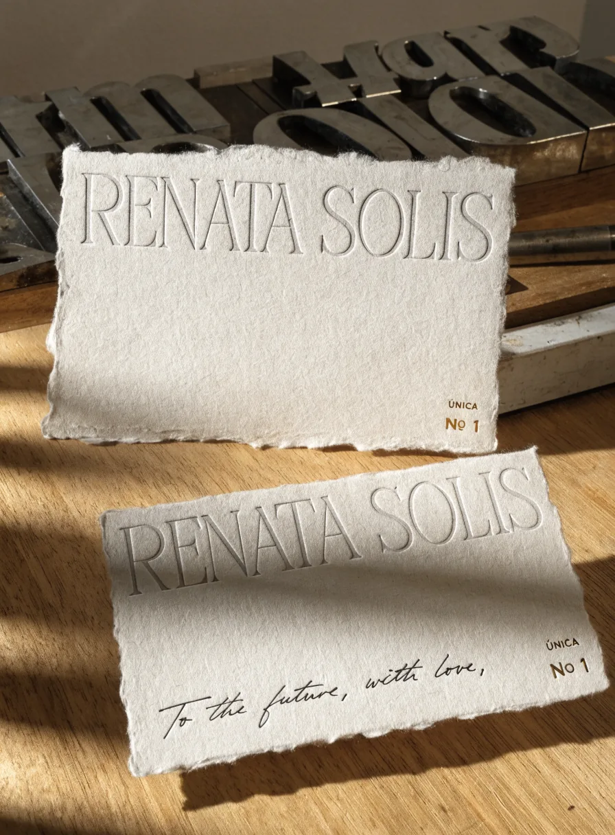 dos tarjetas de papel de algodón hecho a mano con el nombre Renata Solis en relieve al seco, marcadas Única Nº 1 y una dedicatoria manuscrita, sobre un banco de taller tipográfico