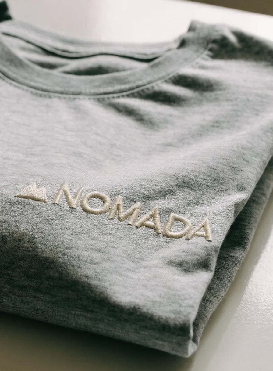 playera de algodón jaspeado gris doblada, con la marca NOMADA bordada al pecho en hilo crema tono sobre tono junto a un pico de montaña