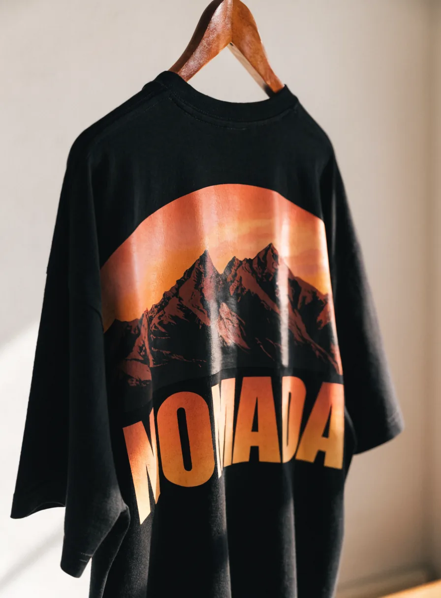 playera oversize negra en un gancho de madera con un paisaje de montañas al atardecer y la palabra NOMADA estampados en grande a la espalda