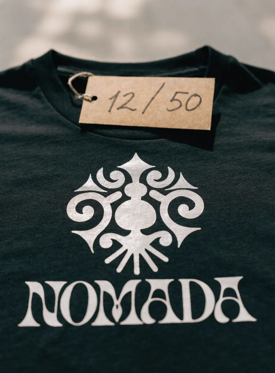 playera verde bosque con un emblema plateado y la palabra NOMADA estampados al centro y una etiqueta de cartón numerada 12 de 50