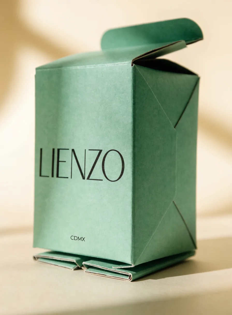 caja plegadiza de cartón color verde salvia con la marca Lienzo impresa en negro y la ciudad CDMX, con la tapa de inserción abierta