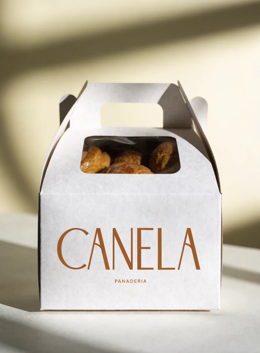 caja plegadiza food-grade con asa y ventana troquelada de la marca Canela Panadería, en cartulina blanca con el nombre en tono terracota, que deja ver el pan dulce adentro