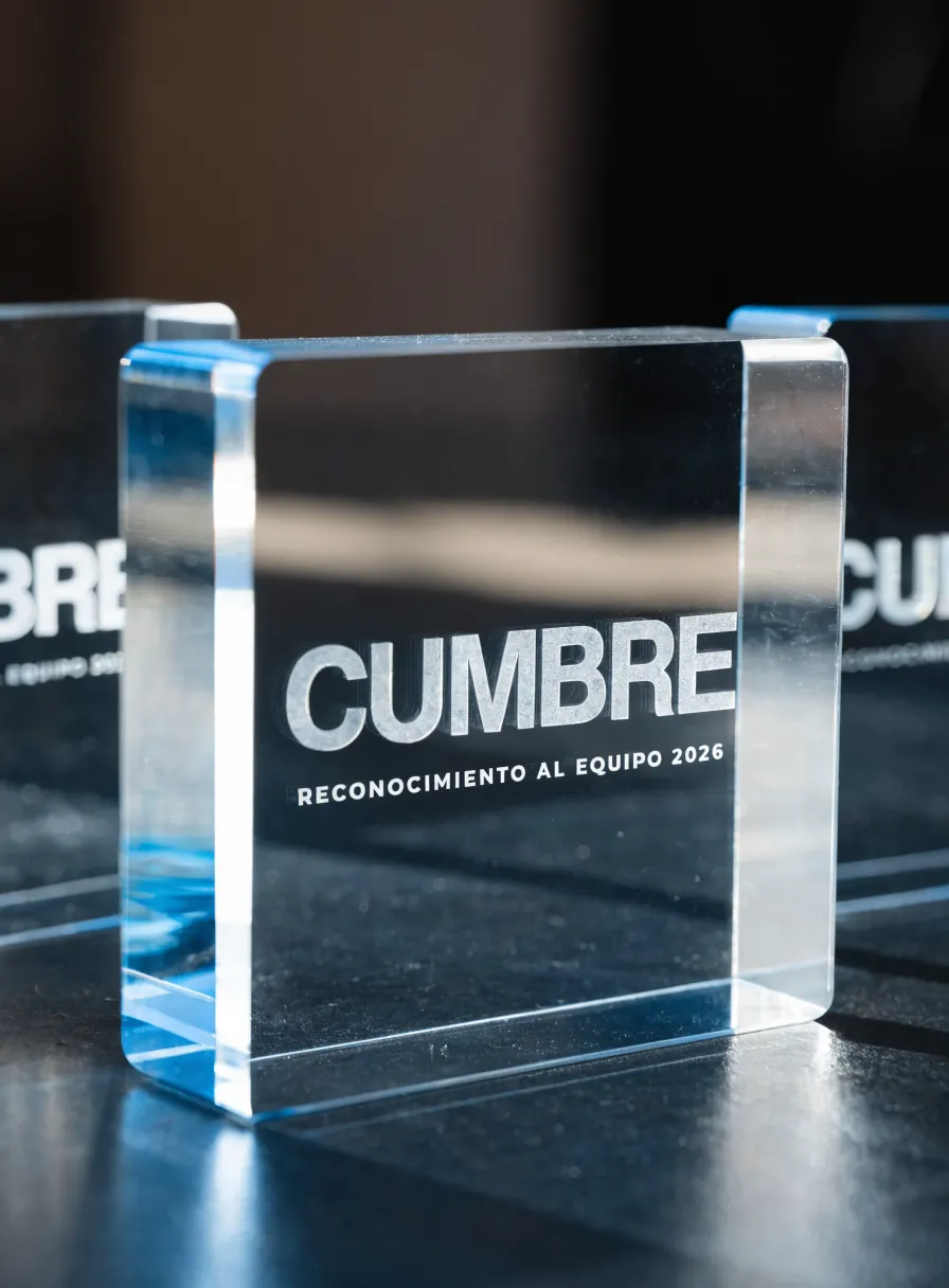 tres reconocimientos de acrílico transparente macizo grabados con la marca ficticia Cumbre y la leyenda Reconocimiento al Equipo 2026, sobre una mesa oscura