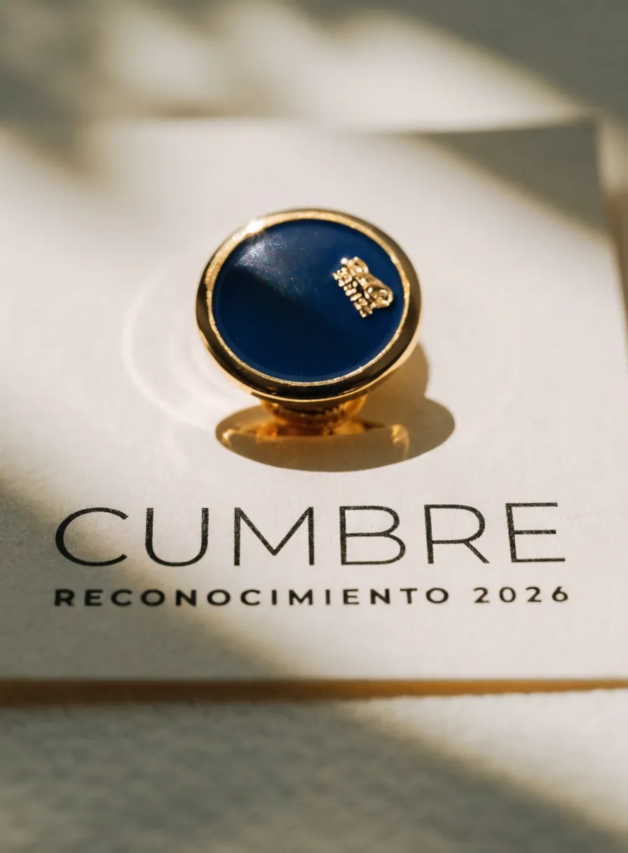 pin de solapa redondo en metal dorado con esmalte azul marino y un pequeño emblema, sobre una tarjeta impresa con la marca ficticia Cumbre Reconocimiento 2026
