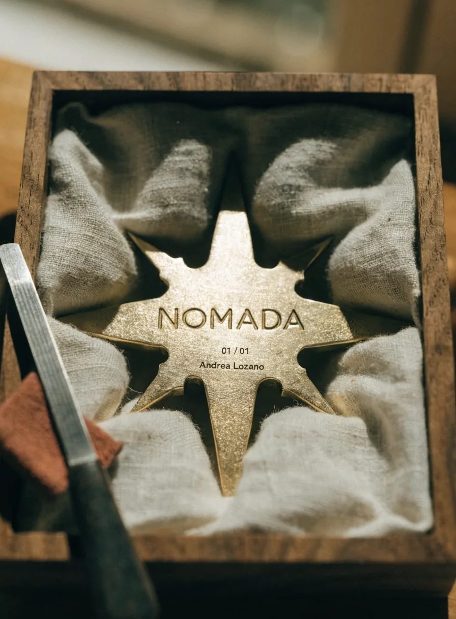 estrella de ocho picos fundida en latón con la palabra NOMADA, la cifra 01/01 y un nombre grabados en la cara, acostada sobre lino crudo dentro de una caja de madera junto a una lima de taller