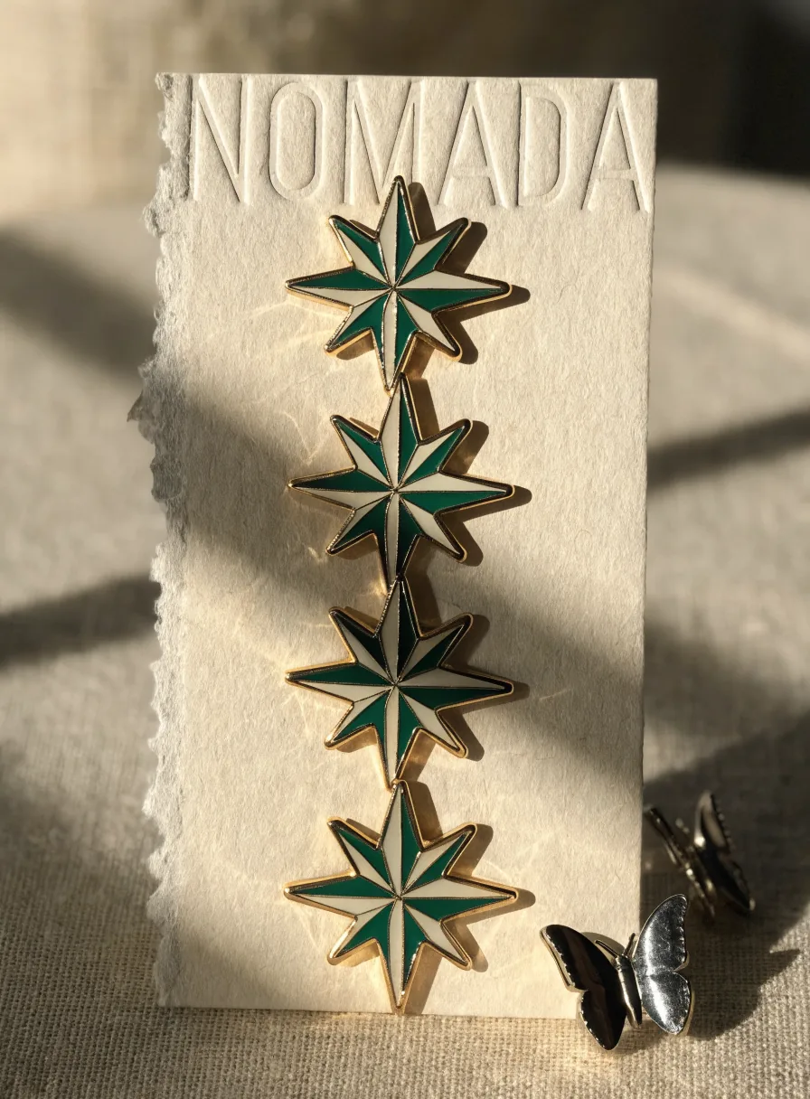 cuatro pines de esmalte duro en forma de rosa de los vientos en verde y crema con baño dorado, prendidos a una tarjeta de algodón con la marca ficticia Nómada hundida en letterpress