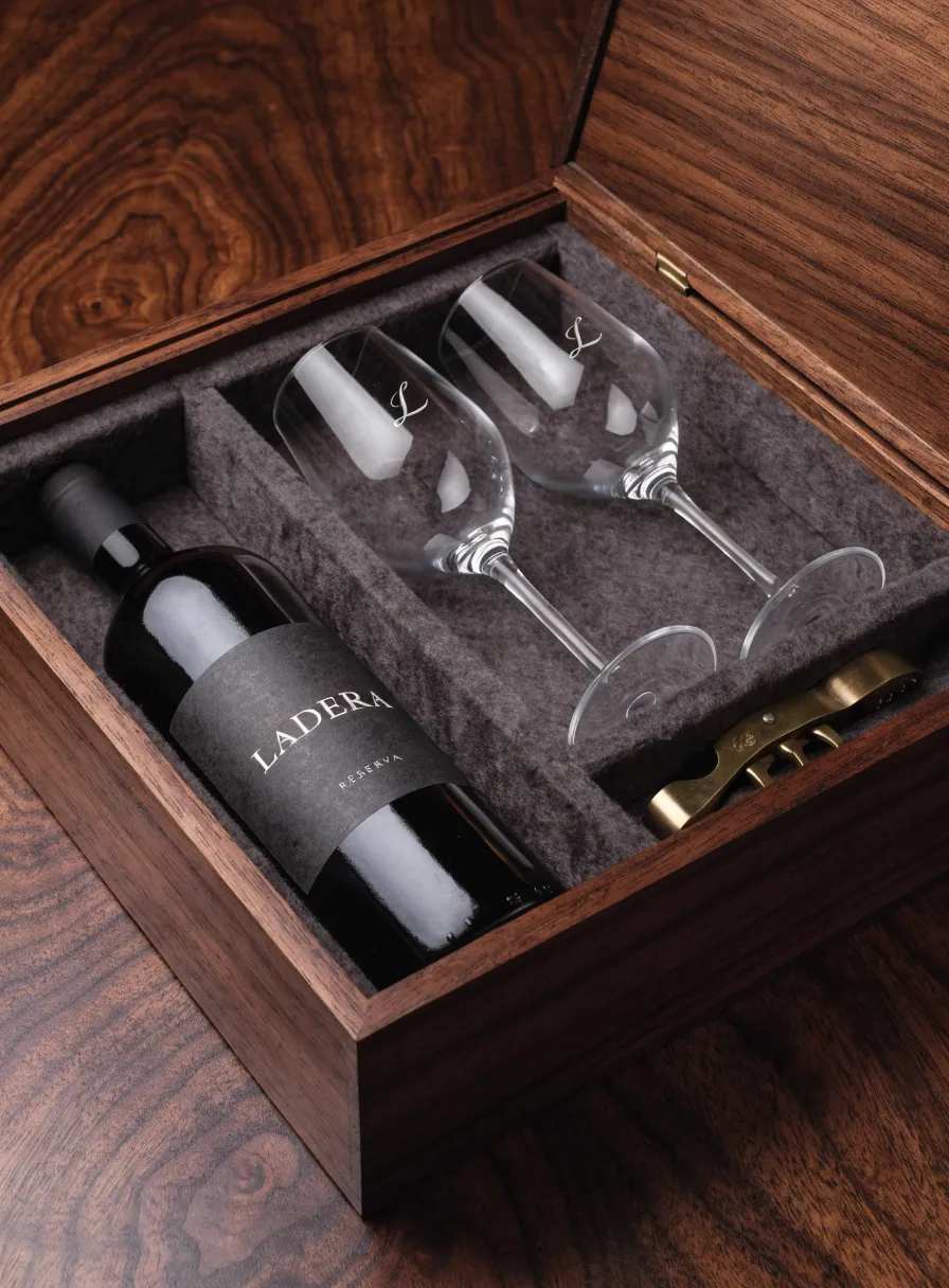 estuche de nogal abierto con una botella de vino Ladera Reserva, dos copas grabadas con el monograma L y un descorchador de latón, sobre forro de fieltro gris
