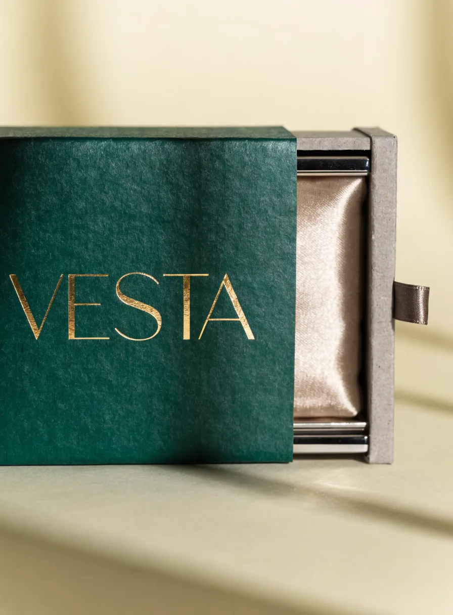 caja cajón rígida en verde esmeralda con la palabra Vesta en foil oro, con la charola interior deslizándose para mostrar un cojín de satín champán y una jaladera de listón