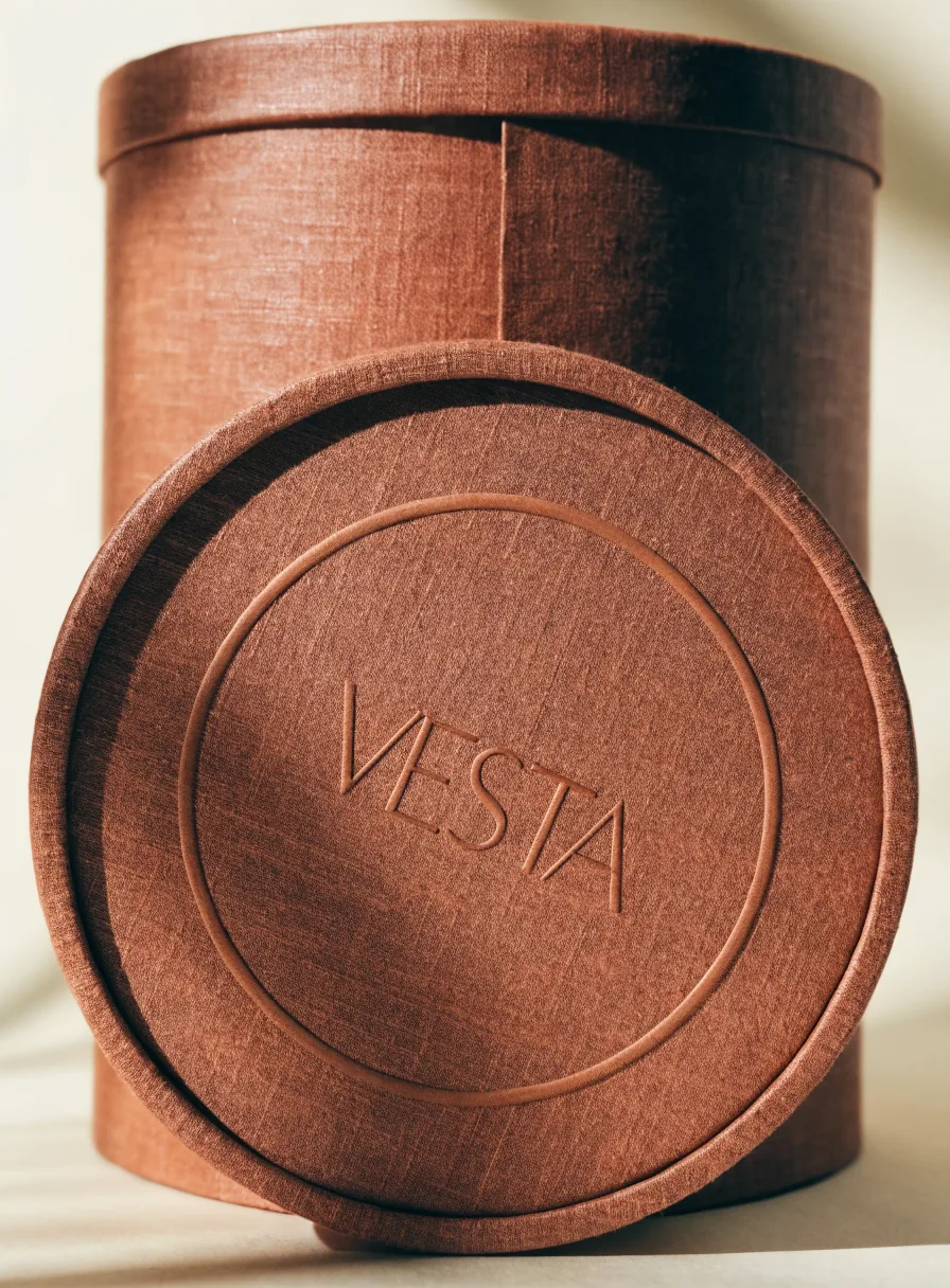 caja rígida cilíndrica forrada en lino color terracota con la palabra Vesta en realce ciego sobre la tapa redonda apoyada al frente