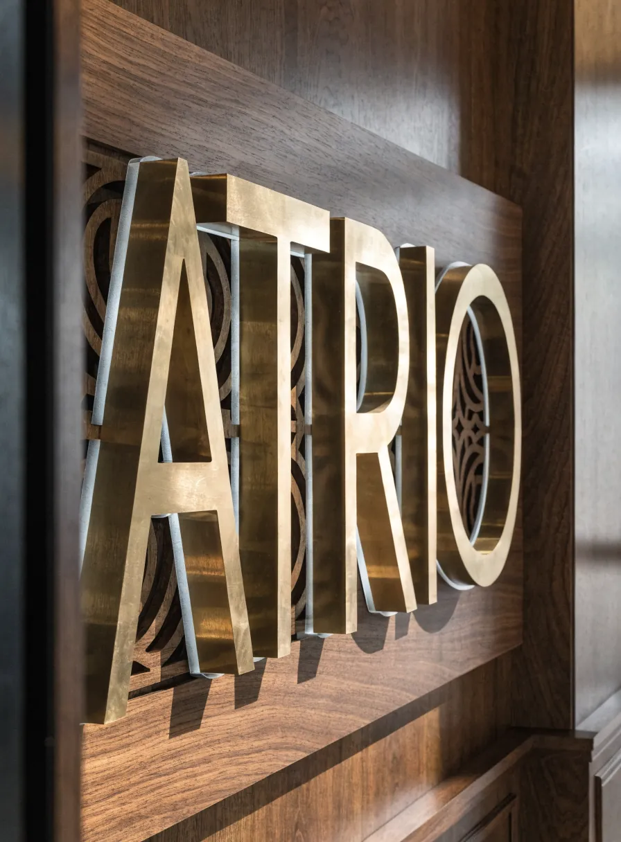 letras dimensionales de latón con la marca ATRIO montadas sobre un muro de nogal con celosía calada al fondo, en una recepción