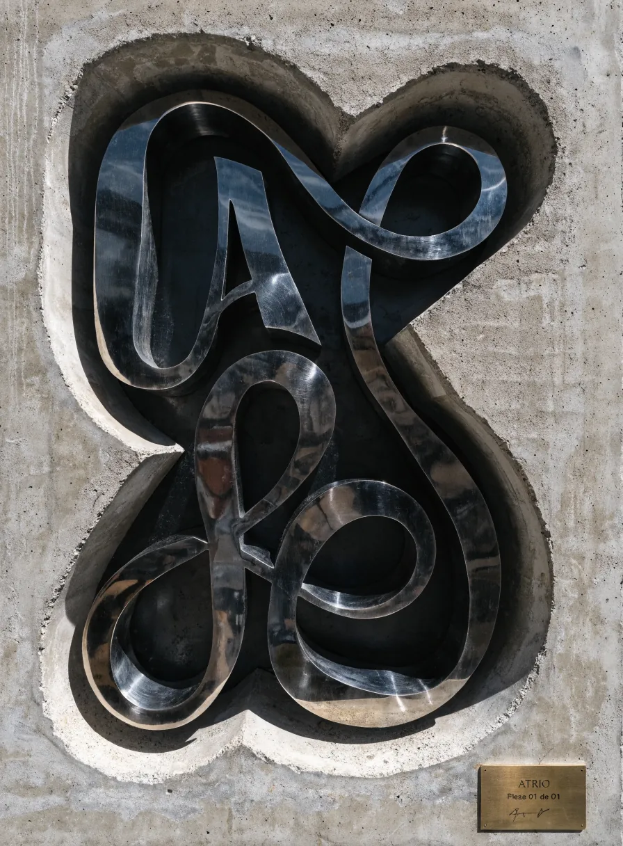 pieza escultórica de letras en metal pulido empotrada en un muro de concreto, con placa de latón que dice ATRIO Pieza 01 de 01 y firma del taller