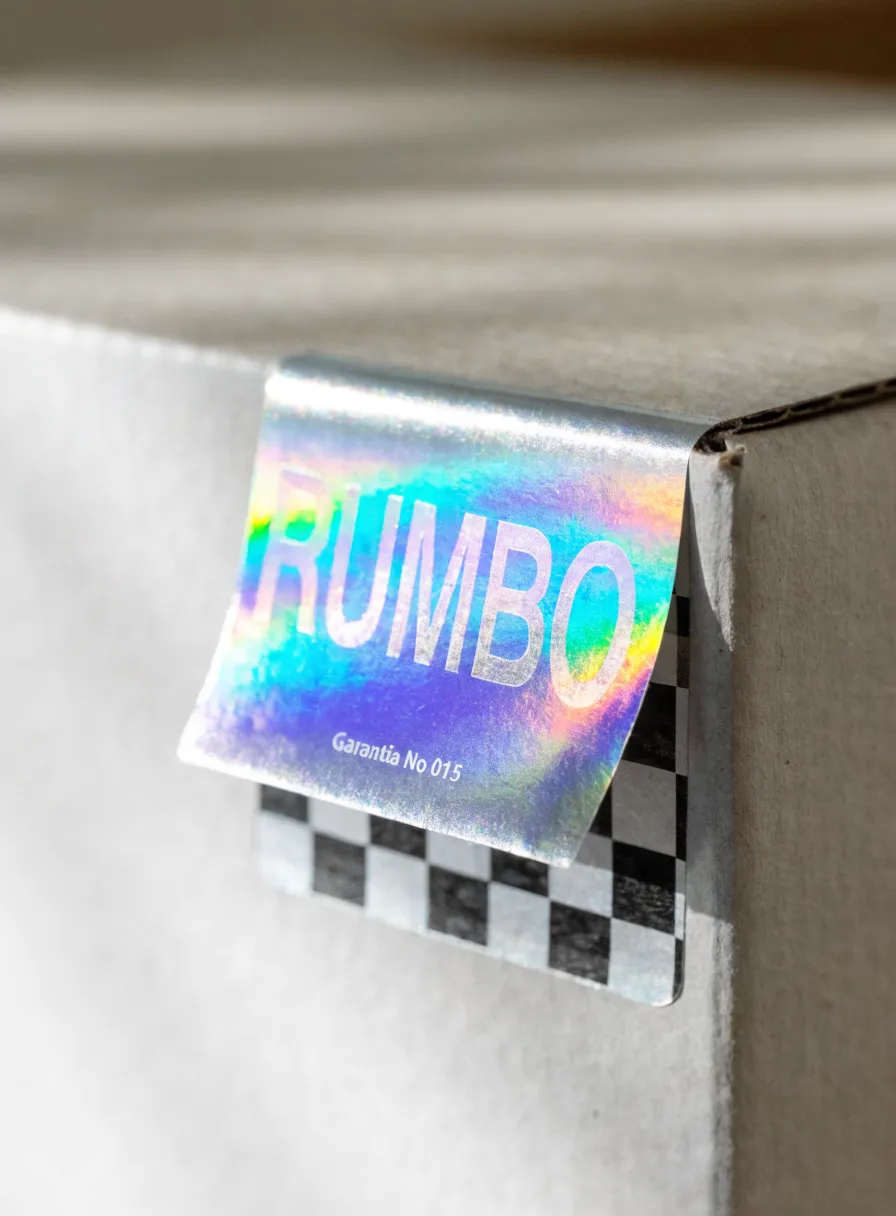 etiqueta de seguridad holográfica despegándose de la esquina de una caja de cartón, con la marca Rumbo en tornasol, el texto Garantía No 015 y un patrón de damero void que queda al levantarla