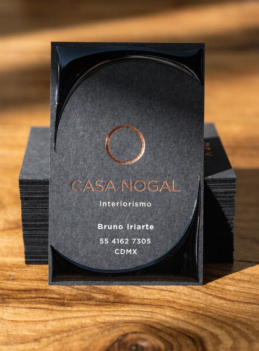 tarjeta vertical de papel negro de la marca ficticia CASA NOGAL, con una curva ancha en barniz UV brillante sobre el papel mate, un aro de foil cobre estampado en la fibra y el nombre en foil cobre, recargada en una pila de tarjetas iguales sobre madera de nogal