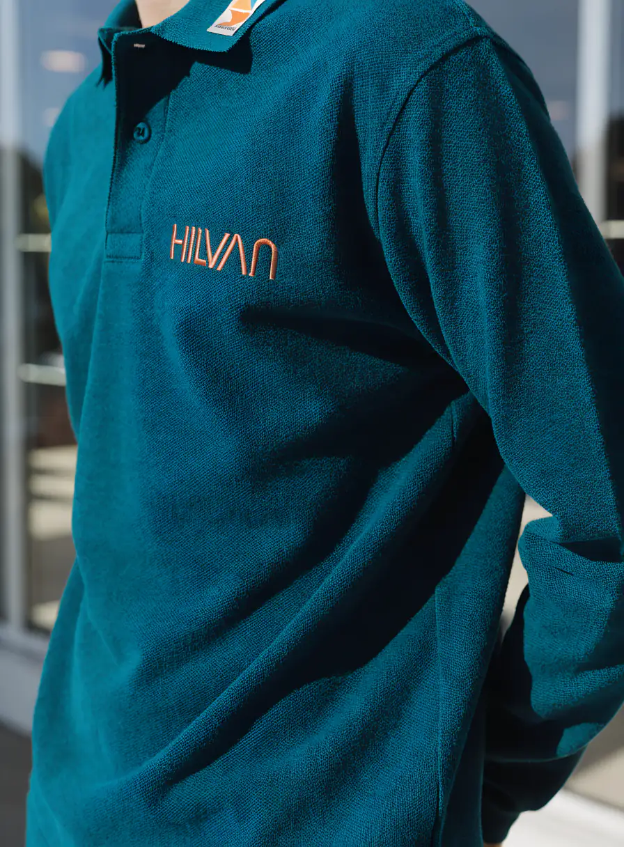 persona con un polo de piqué azul petróleo de manga larga, con la marca HILVAN bordada en hilo naranja sobre el pecho y una etiqueta tejida al cuello