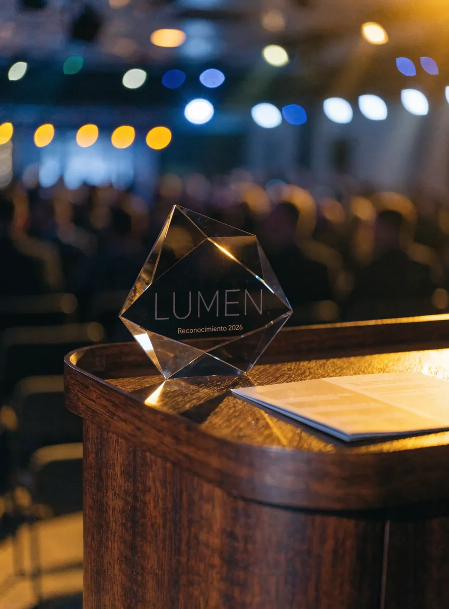 reconocimiento de cristal facetado grabado Lumen Reconocimiento 2026 sobre un atril de madera en el escenario de una ceremonia de premiación, con luces y público al fondo