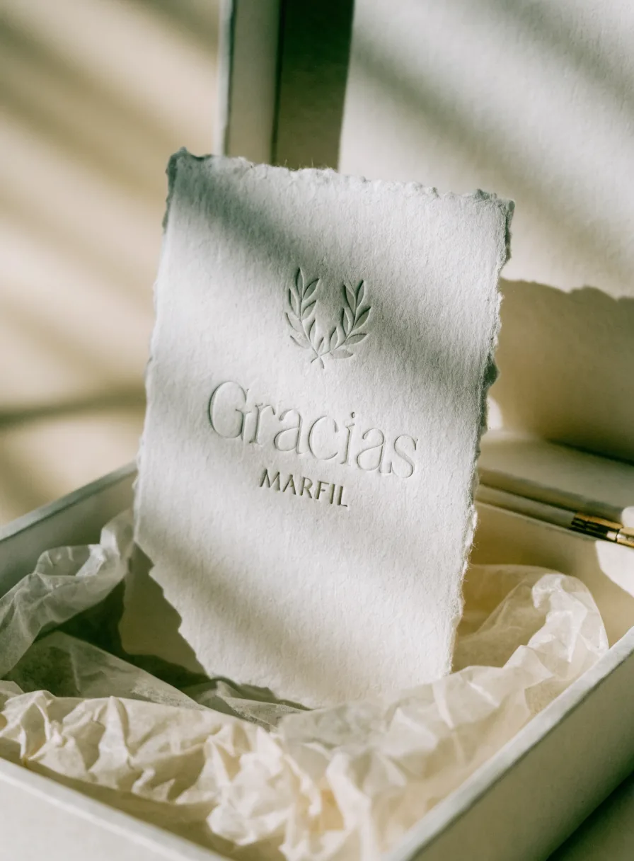 tarjeta de agradecimiento en papel de algodón con canto deckle, con el emblema de laurel y las palabras Gracias y MARFIL en deboss ciego, dentro de una caja con papel china