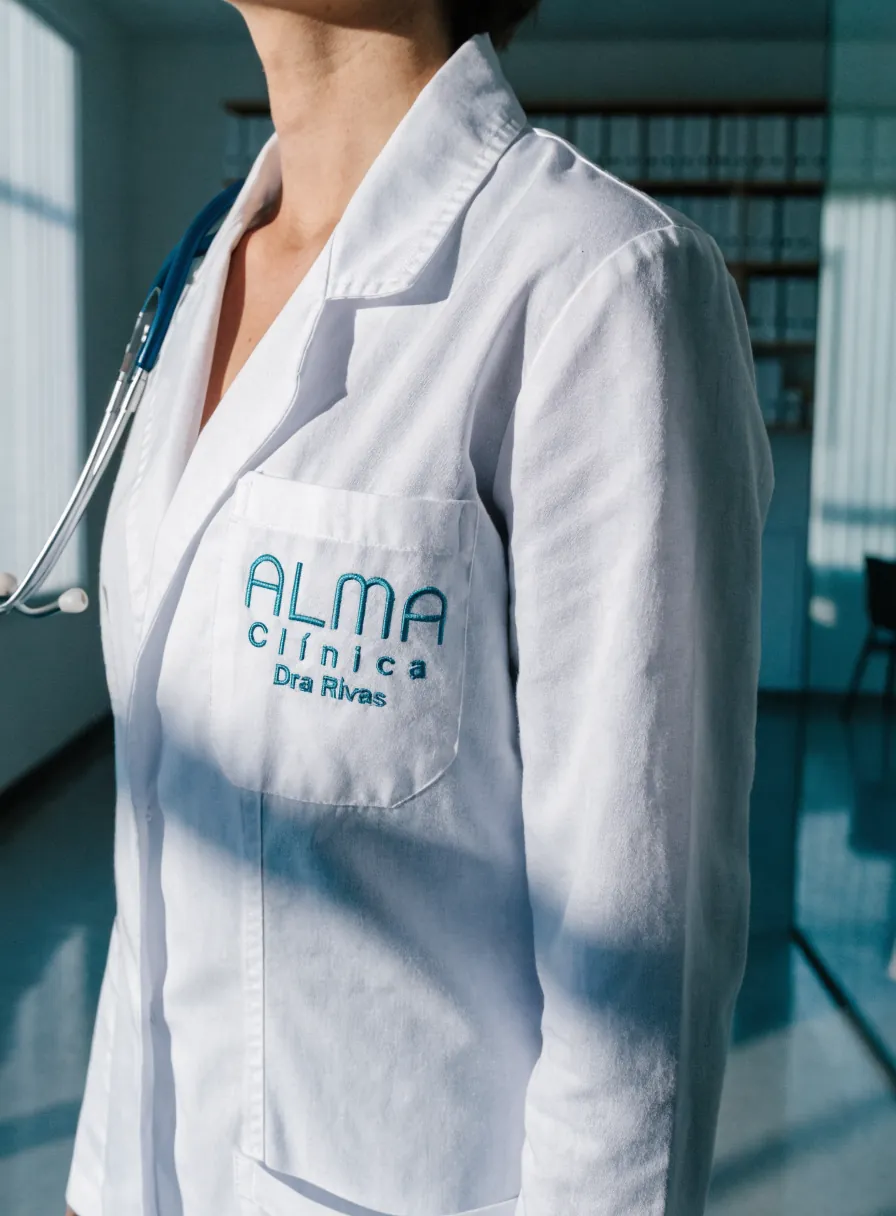 bata clínica blanca puesta con un estetoscopio al cuello, con la marca ficticia Alma Clínica y el nombre Dra Rivas bordados en la bolsa del pecho