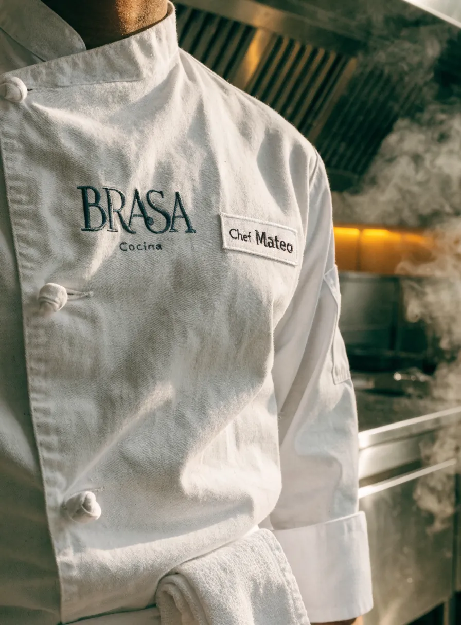 filipina de chef blanca de doble botonadura puesta en una cocina, con la marca ficticia Brasa Cocina bordada al pecho y un parche de nombre que dice Chef Mateo