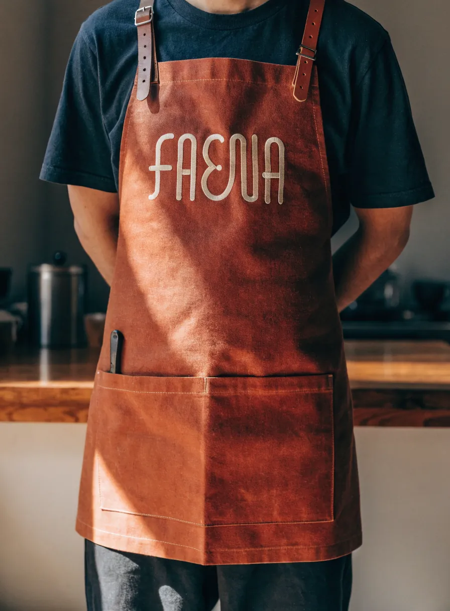 mandil de lona encerada color terracota con correas de piel puesto por un barista, con la marca ficticia Faena al frente y una bolsa canguro