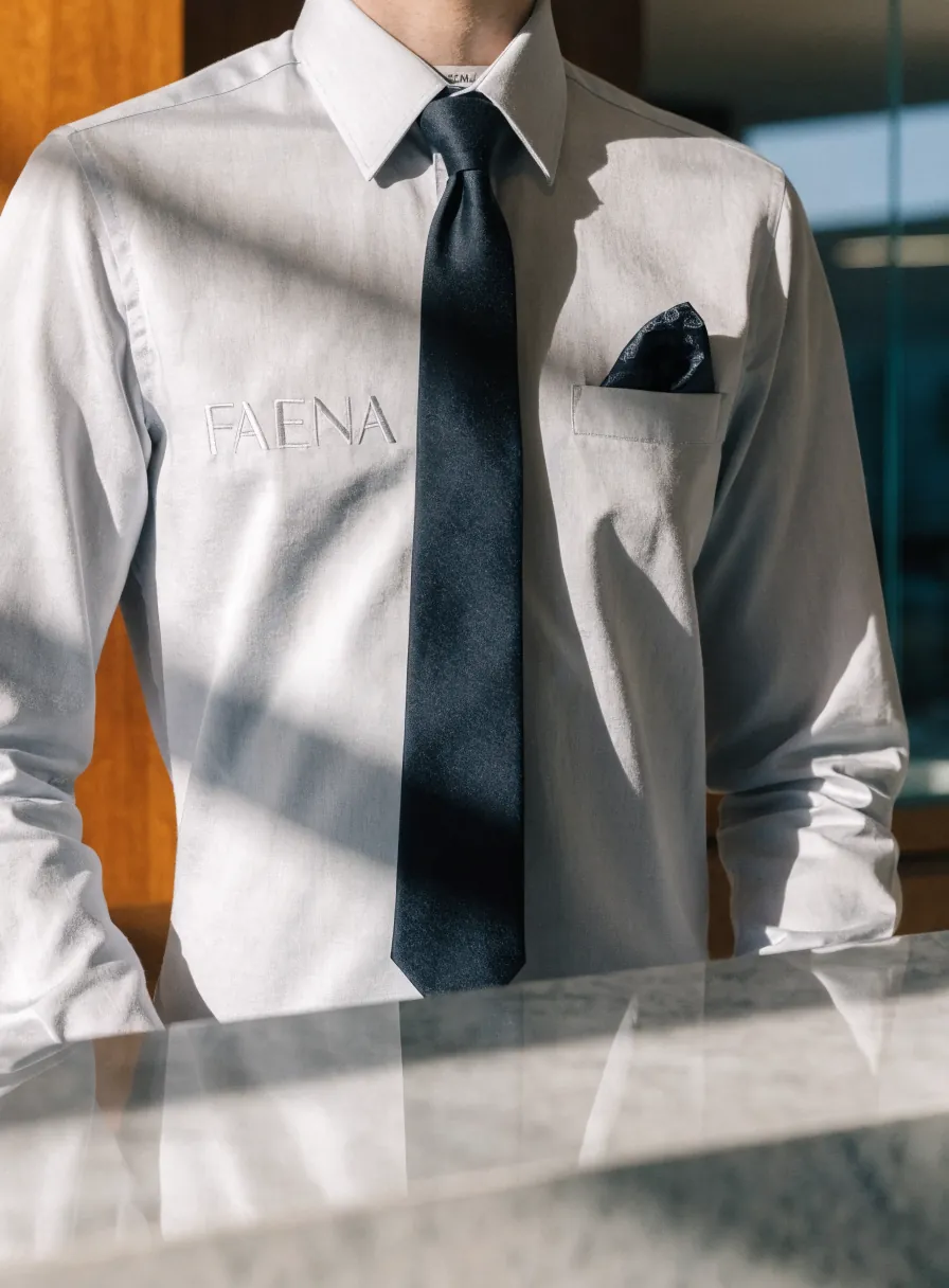 camisa de vestir blanca con corbata azul marino y pañuelo al bolsillo puesta en una recepción, con la marca ficticia Faena bordada tono sobre tono al pecho