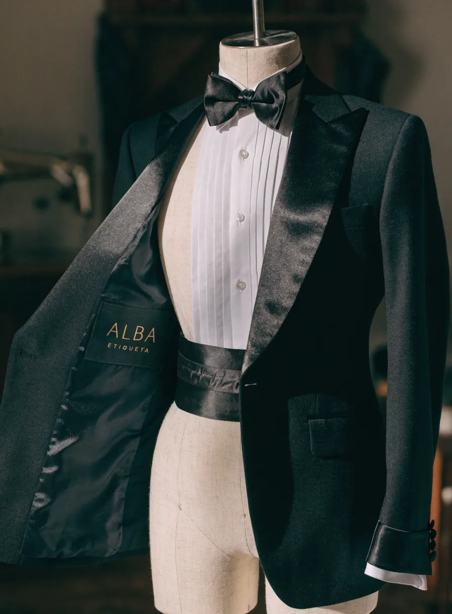 esmoquin negro sobre un maniquí con solapa de seda, camisa de plastrón plisado, corbatín de moño, faja y forro con la etiqueta ALBA