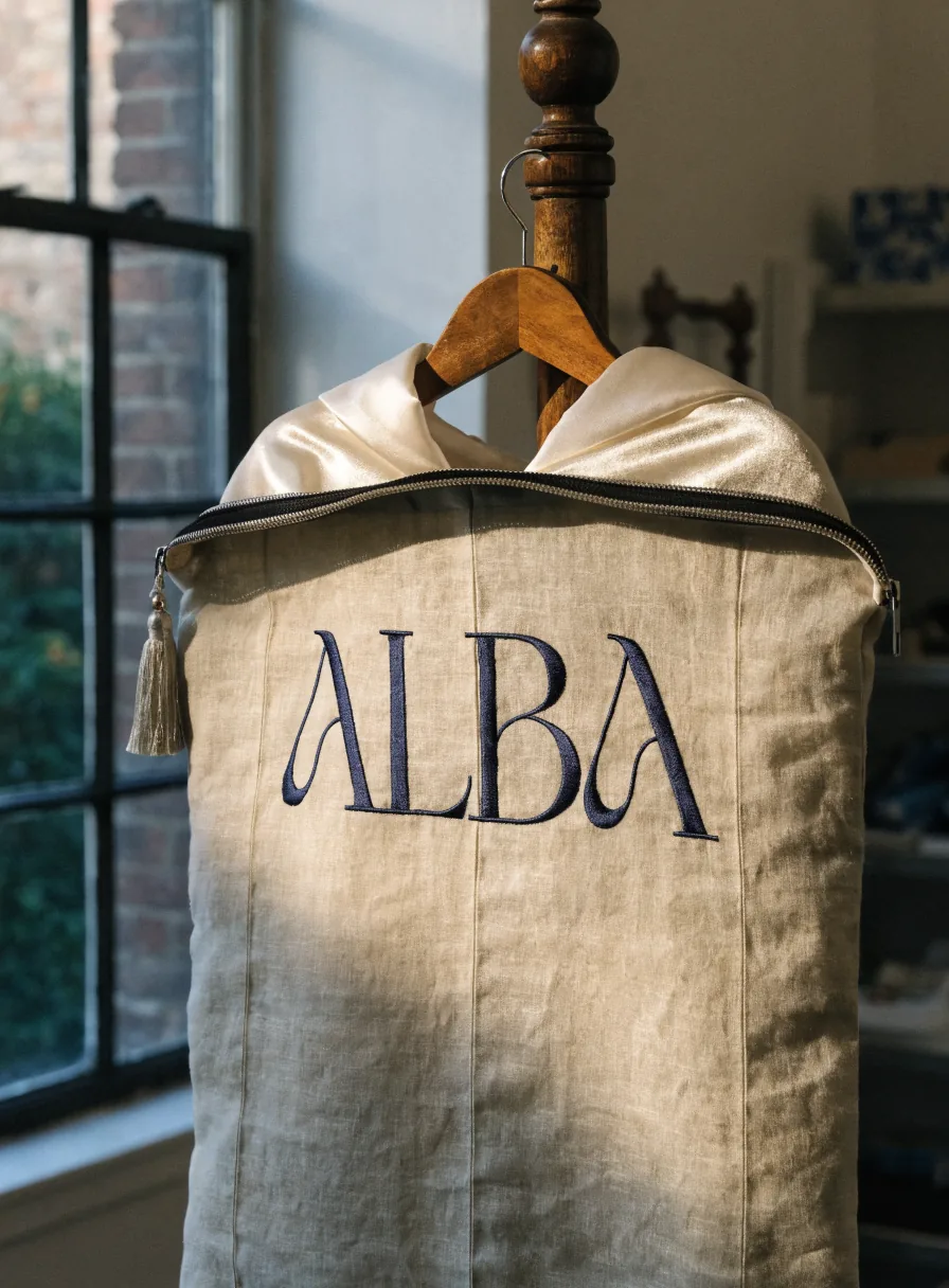 funda de vestido de lino natural con la palabra ALBA bordada al frente, colgada en un burro de madera con la cremallera entreabierta que deja ver el hombro de raso del vestido dentro, y un cordón con borla