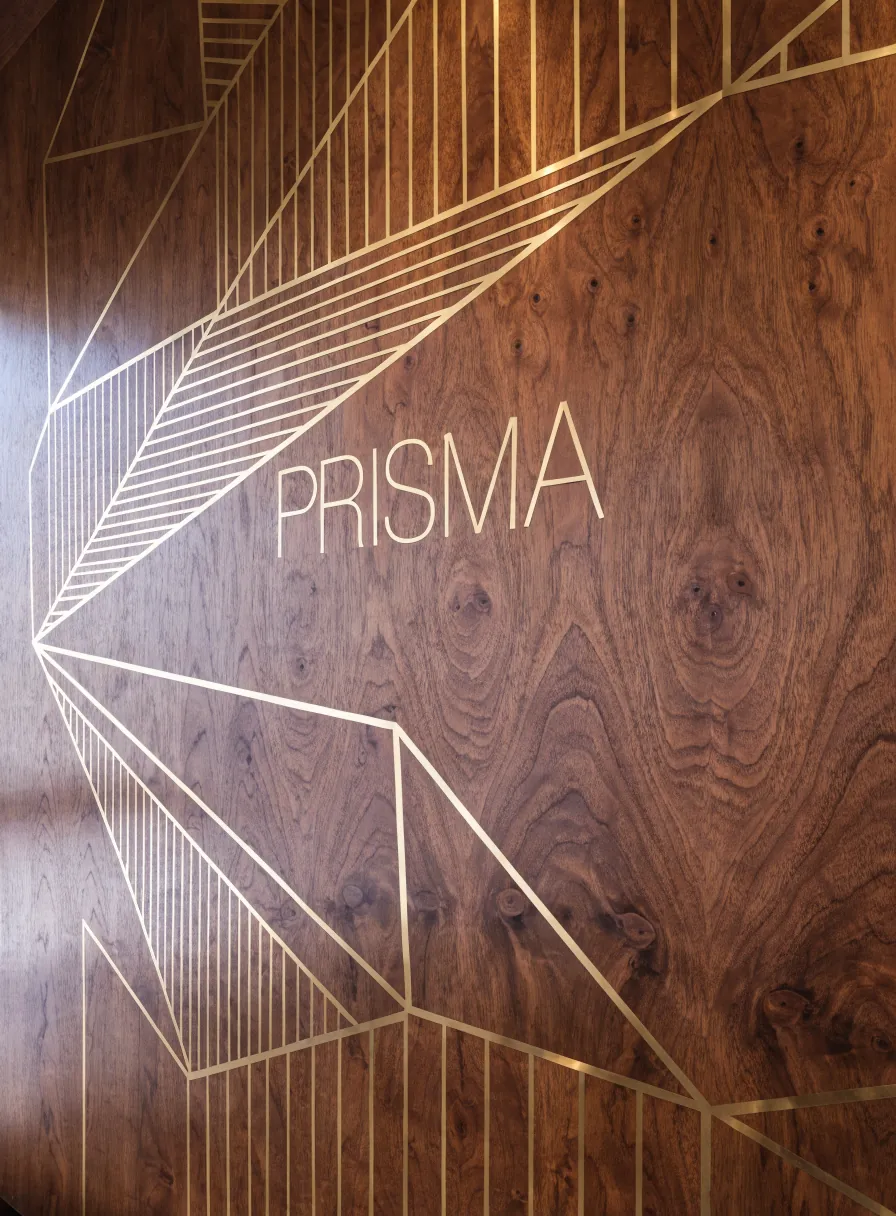 muro de madera de nogal con líneas finas de latón incrustadas que irradian en abanico y el nombre PRISMA en latón al centro