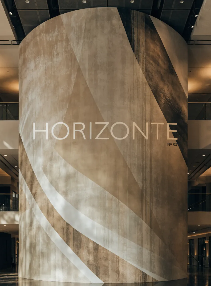 columna curva forrada con un envolvente de líneas geométricas en café y beige, la marca Horizonte número 02, en el vestíbulo de piso pulido de un edificio con barandales de vidrio