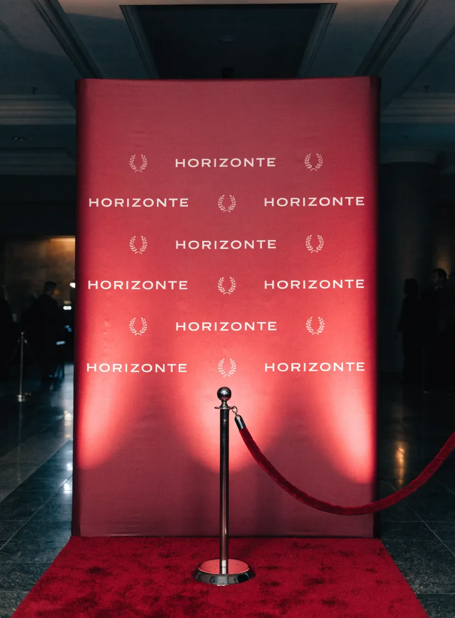 backdrop rojo con la marca Horizonte repetida y un emblema de laurel, montado en un vestíbulo con alfombra roja y cordón de terciopelo