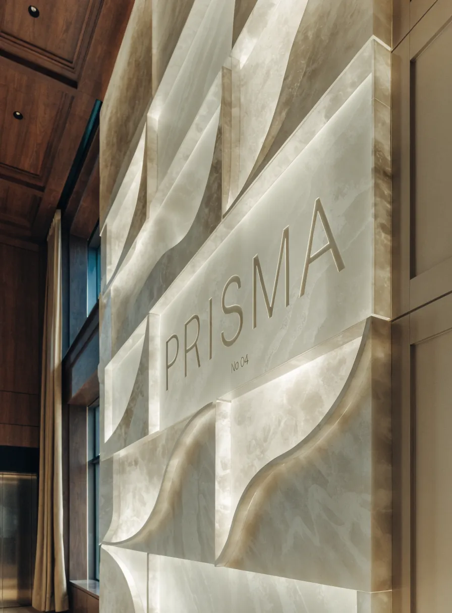 mural de paneles tallados en piedra clara con formas onduladas, retroiluminado desde cada junta y con canto de latón, el nombre de la marca ficticia PRISMA grabado en el panel central, en un pasillo corporativo con paneles de madera