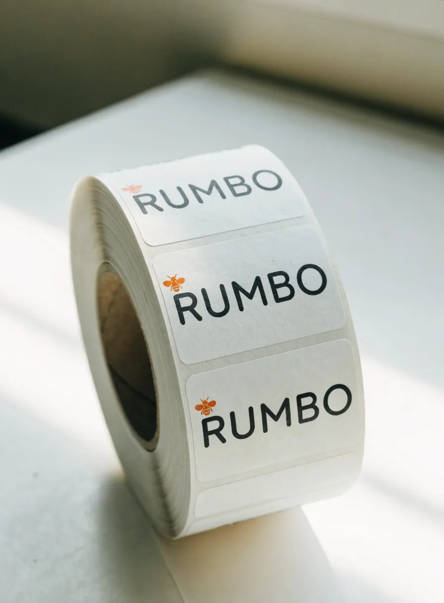 rollo de etiquetas de papel adhesivo con la marca Rumbo y una abeja en naranja, sobre una repisa junto a la ventana