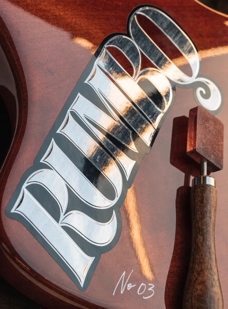 transfer metálico plateado con la palabra Rumbo en letras ornamentadas, bruñido a mano sobre el cuerpo de una guitarra de madera, con la espátula de bruñido al lado y el número 03