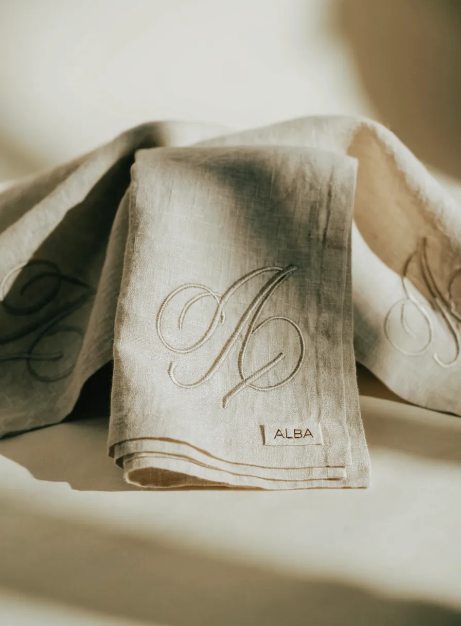 Servilletas de lino natural dobladas con un monograma bordado en cursiva y la marca ALBA bordada en la esquina