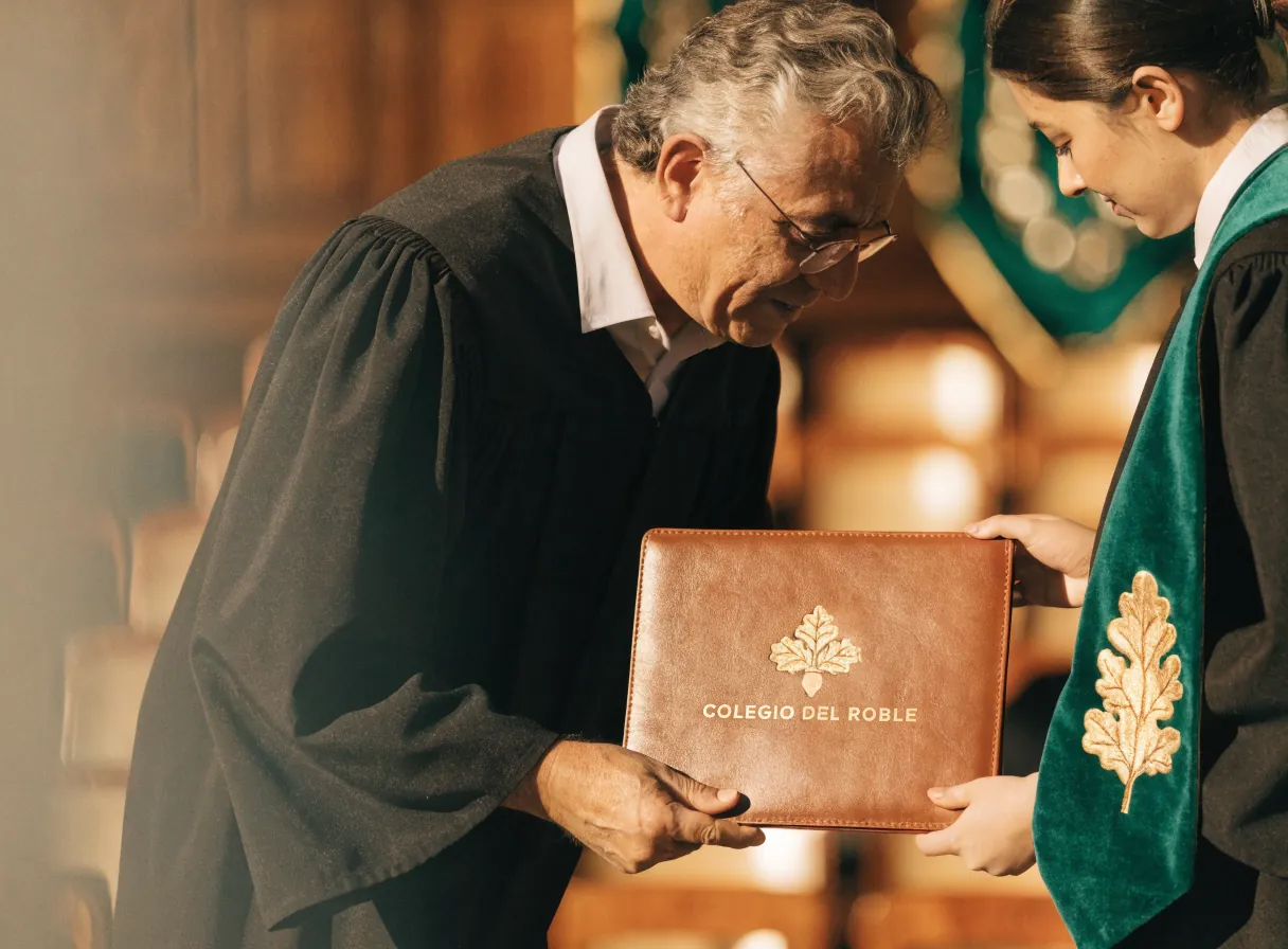 el rector entrega un portafolio de diploma de piel con el escudo en foil oro a una graduada con estola verde y la hoja de roble bordada en hilo de oro, en la ceremonia del Colegio del Roble