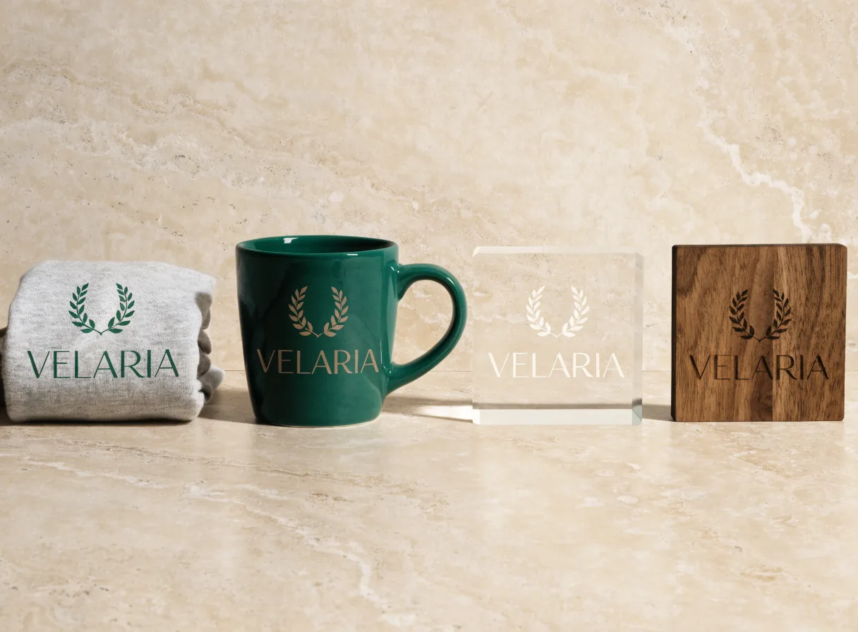 El mismo monograma VELARIA en cuatro materiales, con serigrafía sobre una playera de algodón, esmalte sobre una taza de cerámica verde, grabado interior en un bloque de cristal y láser sobre un posavasos de nogal